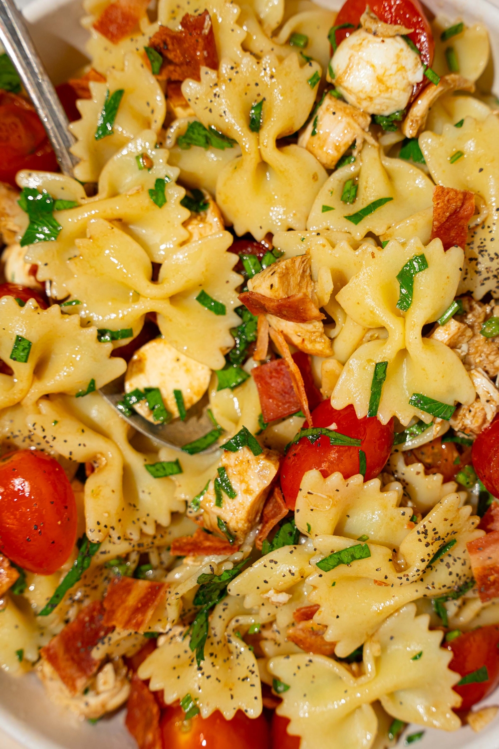 A white bowl of bruschetta chicken pasta with bacon tossed with bowtie pasta, mozzarella cheese, tomatoes, and basil in a balsamic vinaigrette. There is a fork on the bowl.