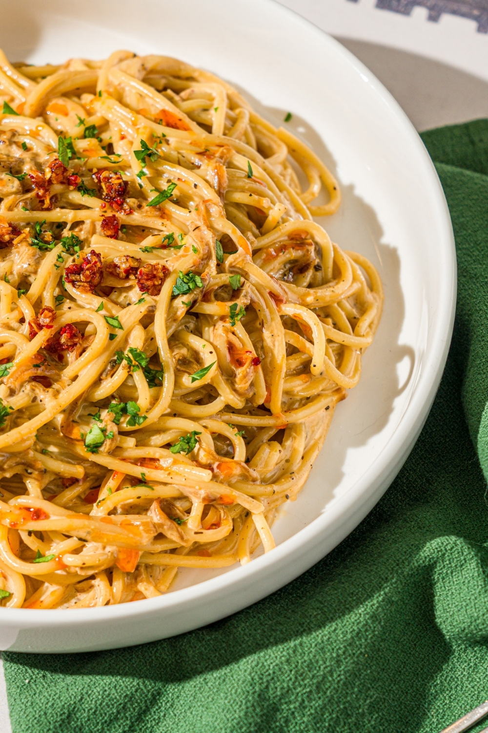 A white bowl with caramelized onion pasta garnished with chili crisp onion and parsley. The bowl is on a tiled counter with a green cloth napkin.