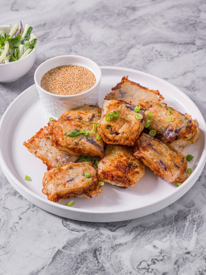 A white plate with chicken and shrimp rice paper dumplings garnished with sliced green onions and served with a small bowl of dipping sauce. The plate is on a marble counter with a small side salad in a bowl.
