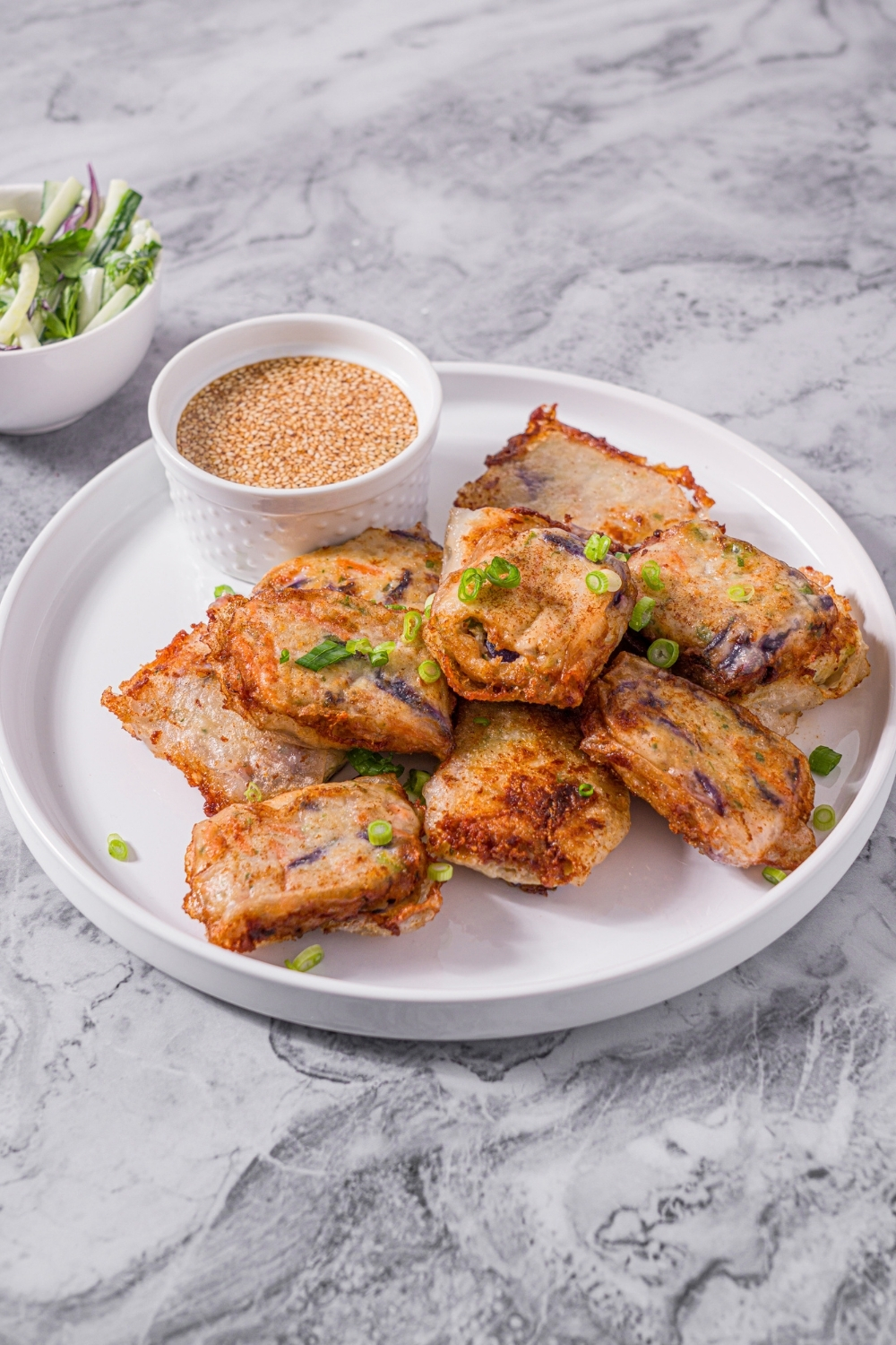 A white plate with chicken and shrimp rice paper dumplings garnished with sliced green onions and served with a small bowl of dipping sauce. The plate is on a marble counter with a small side salad in a bowl.