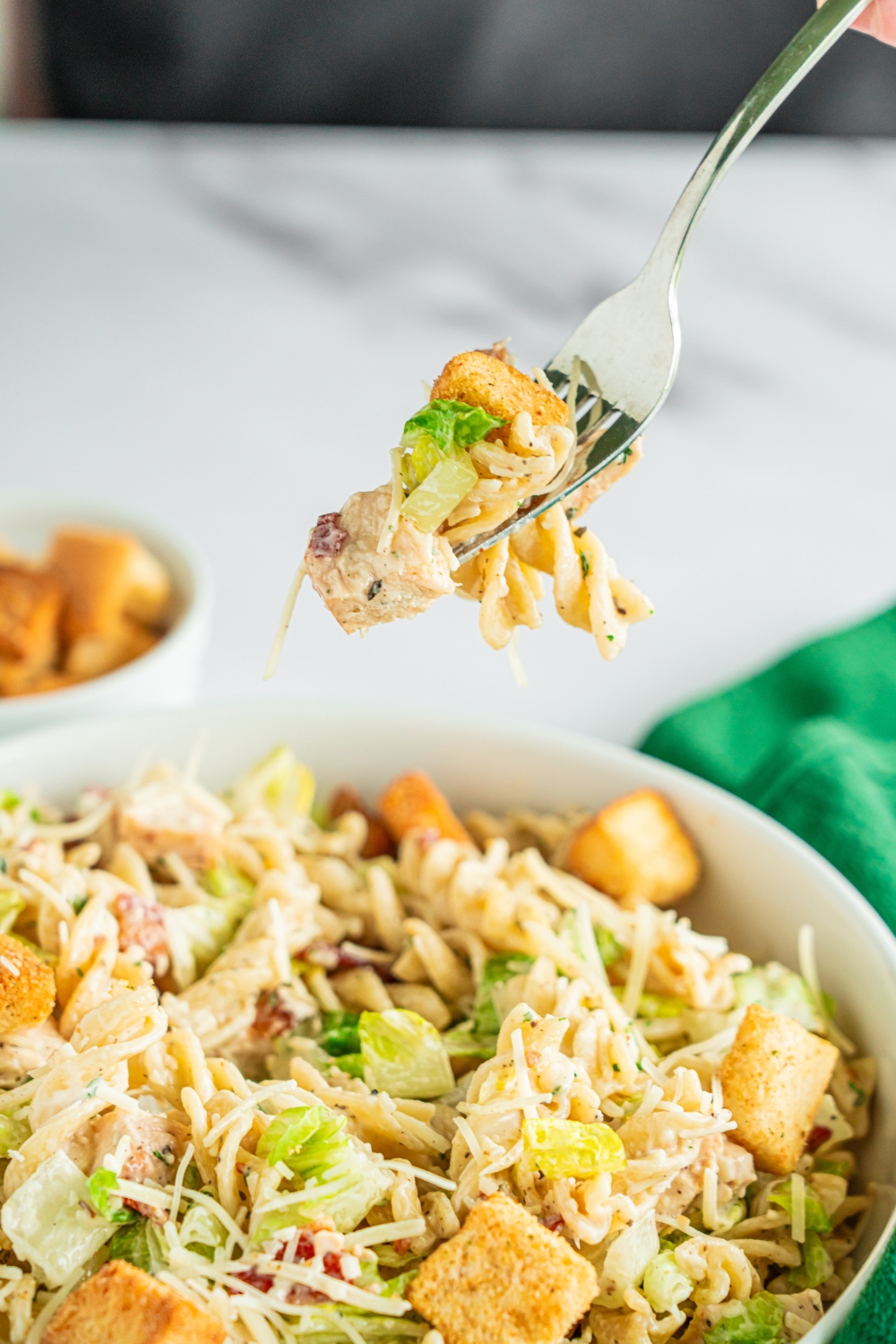 A fork with a bite of chicken caesar pasta salad over a bowl of pasta salad topped with shredded parmesan cheese and croutons. The bowl is on a marble counter with a green cloth napkin and small bowl of croutons.