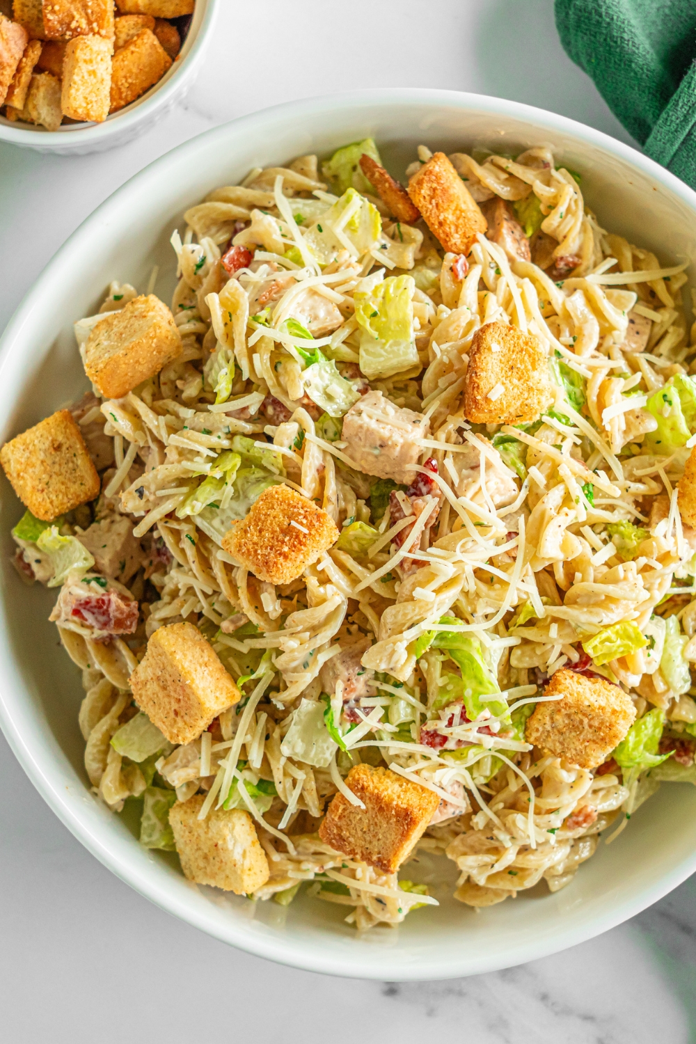 A white bowl of chicken caesar pasta salad topped with shredded parmesan cheese and croutons. The bowl is on a marble counter with a green cloth napkin and small bowl of croutons.