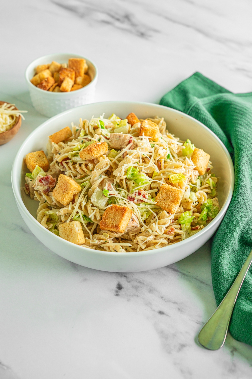 A white bowl of chicken caesar pasta salad topped with shredded parmesan cheese and croutons. The bowl is on a marble counter with a green cloth napkin, fork, and small bowl of croutons.
