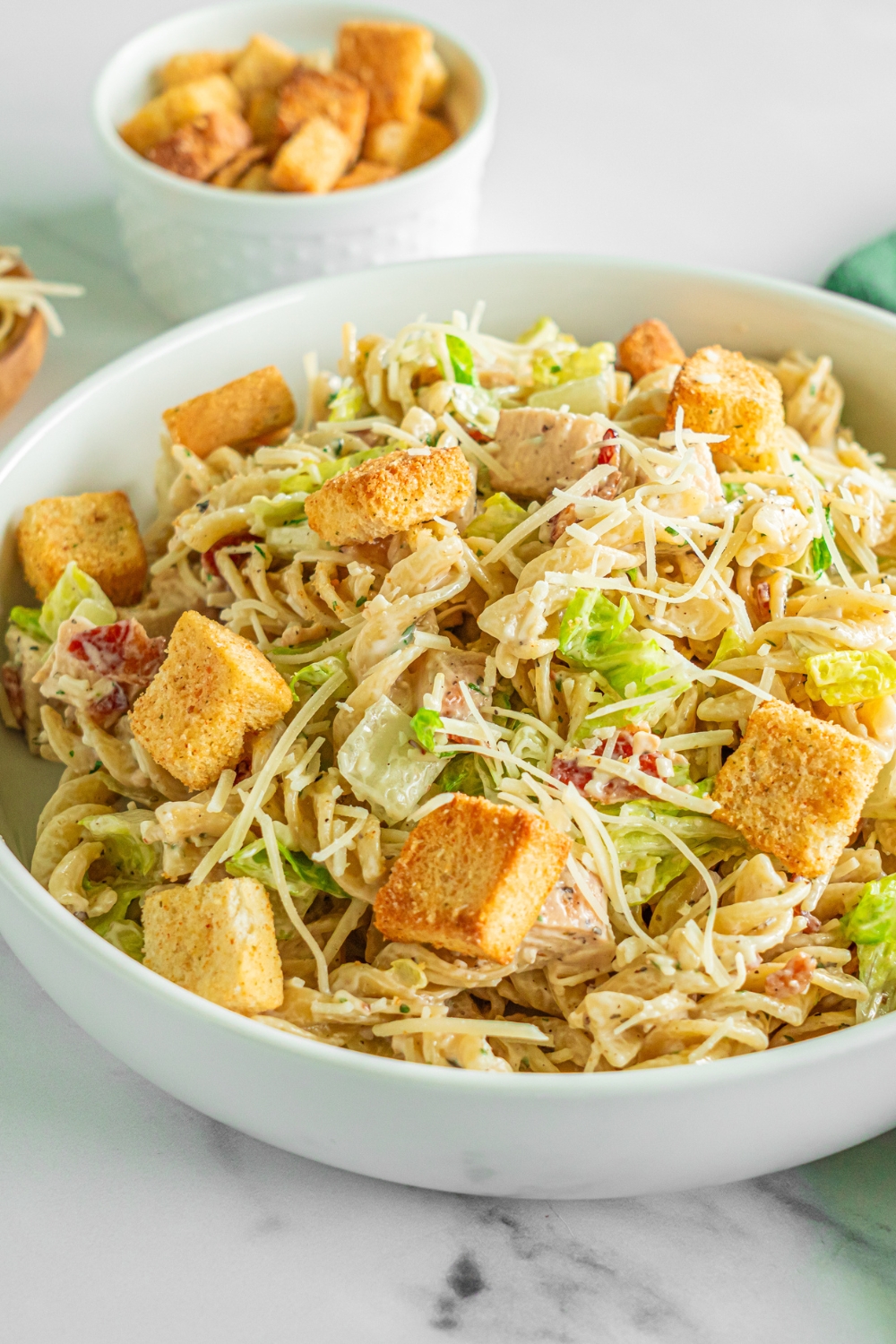A white bowl of chicken caesar pasta salad topped with shredded parmesan cheese and croutons. The bowl is on a marble counter with a green cloth napkin and small bowl of croutons.