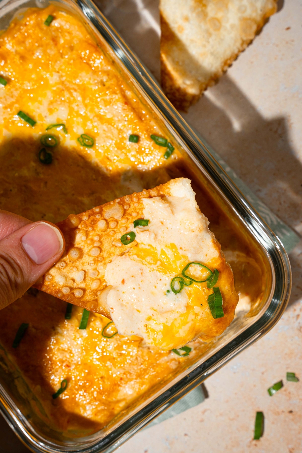 A glass baking dish with baked crab rangoon dip garnished with sliced green onions. A hand is dipping a wonton chip into the dip. The dish is on a tan counter.
