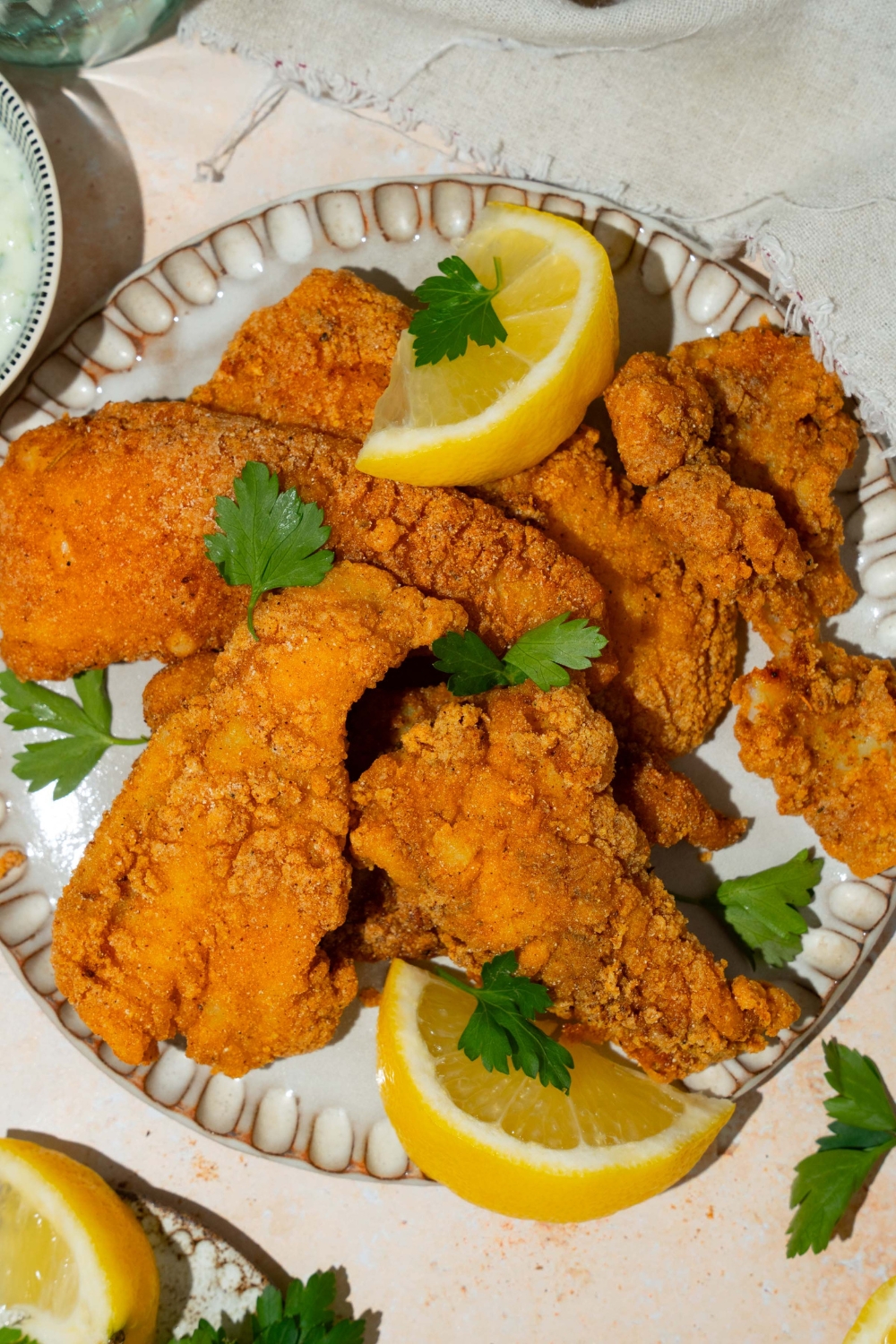 A white plate with several pieces of crispy Southern style fried fish garnished with fresh parsley and served with lemon. The plate is on a tan counter with a white cloth napkin.