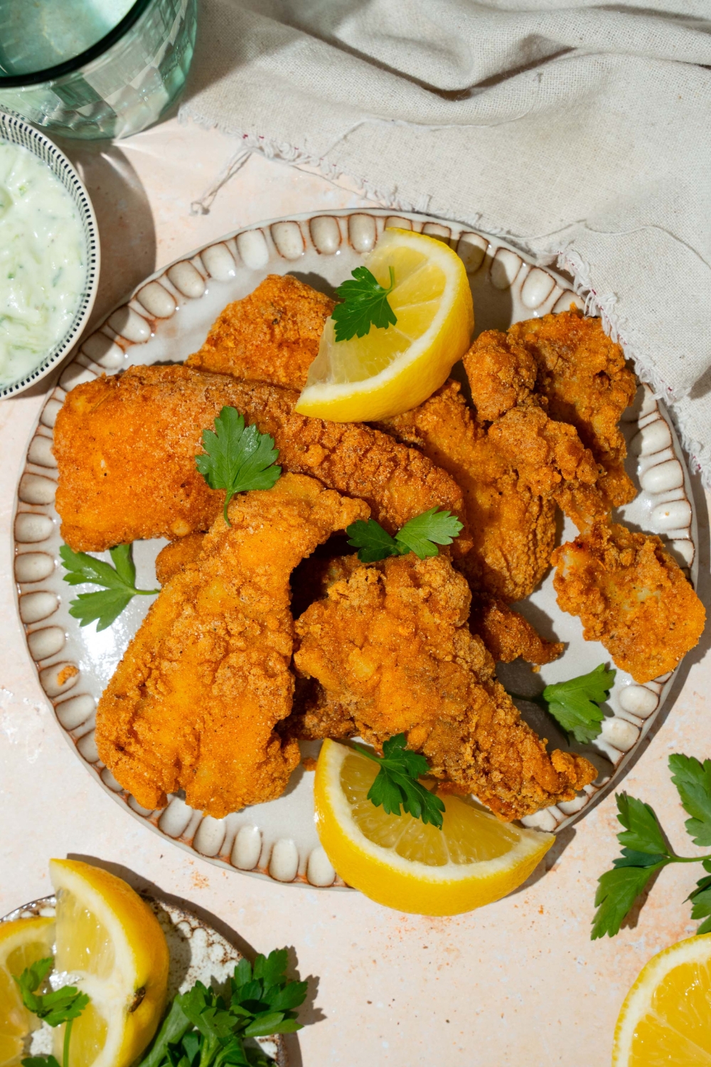 A white plate with several pieces of crispy Southern style fried fish garnished with fresh parsley and served with lemon. The plate is on a tan counter with a white cloth napkin.