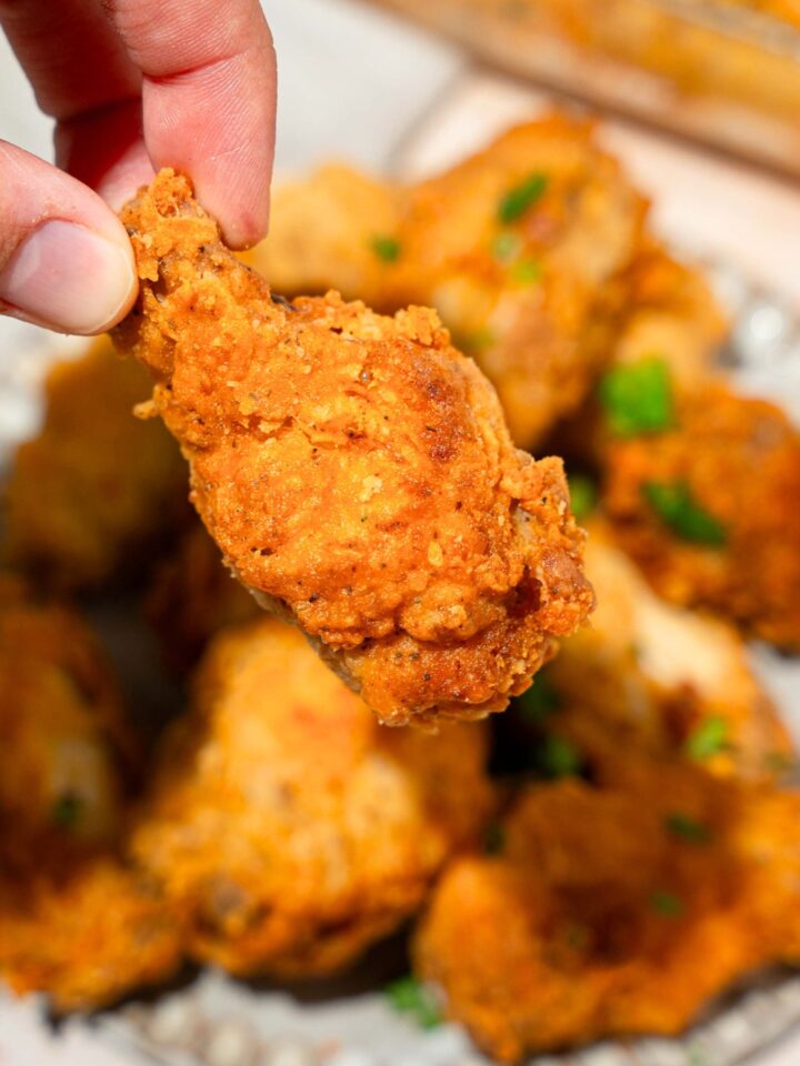 Fingers holding a crispy fried breaded chicken wing over a plate with more fried breaded chicken wings on it.