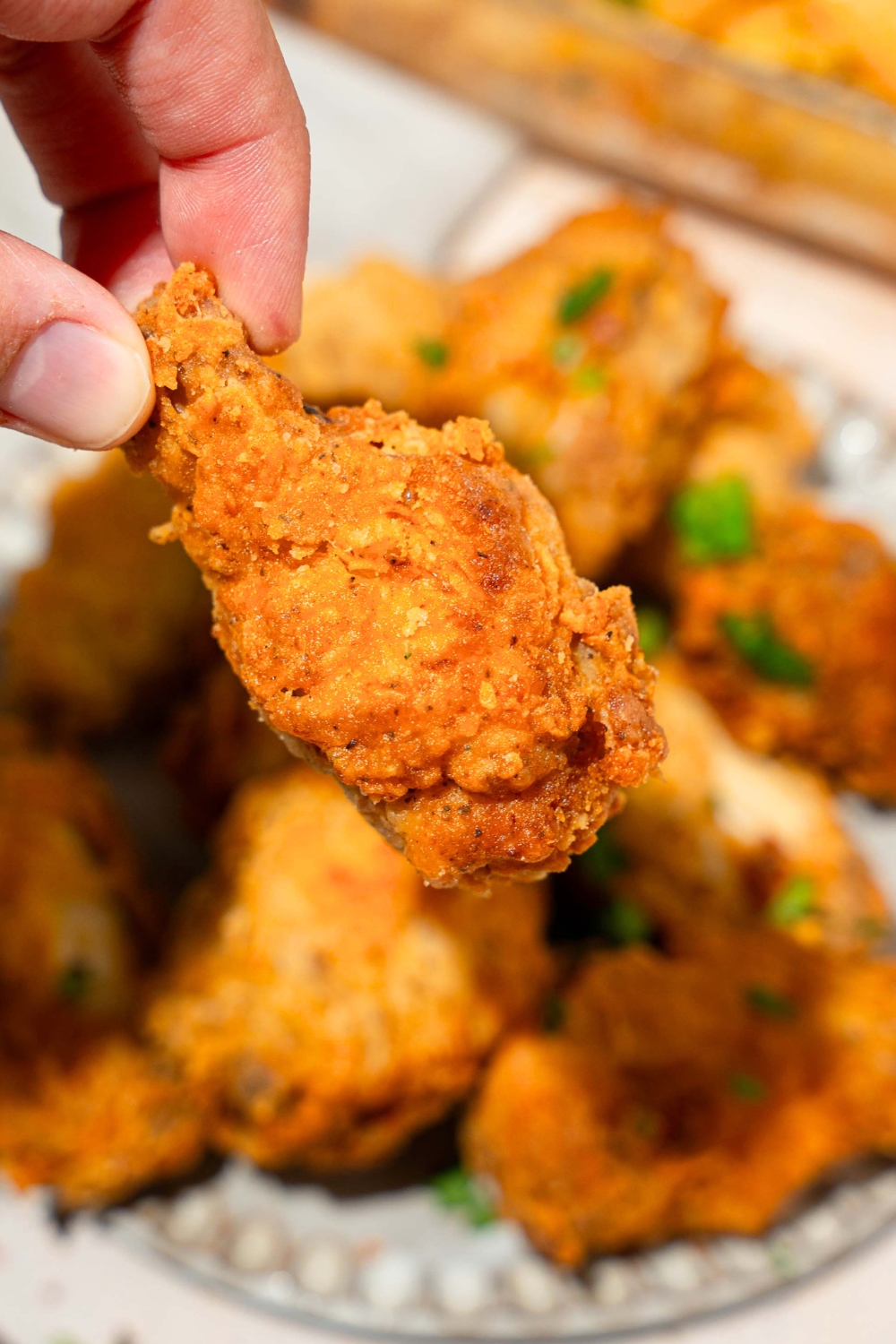 Fingers holding a crispy fried breaded chicken wing over a plate with more fried breaded chicken wings on it.