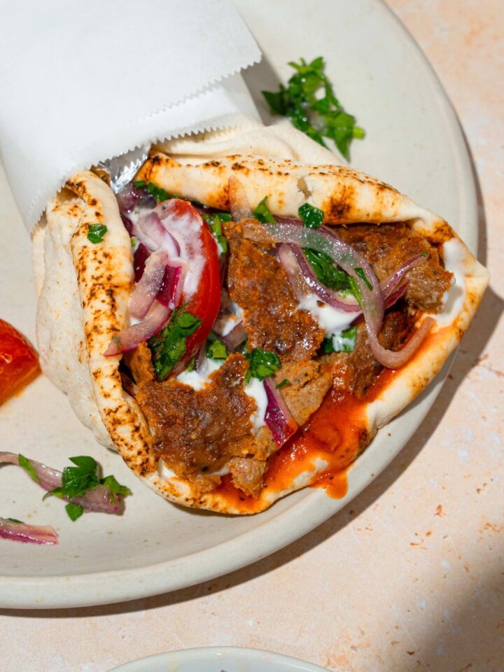 A doner kebab with the beef. onions, tomato, and sauce wrapped in a pita that's wrapped in paper and set on a white plate.