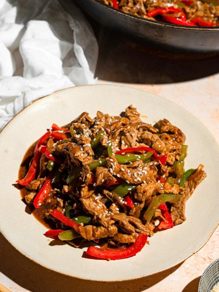 A bunch of pieces of sliced flank steak and red and green bell peppers and onions coated in a stir fry sauce on a white plate.