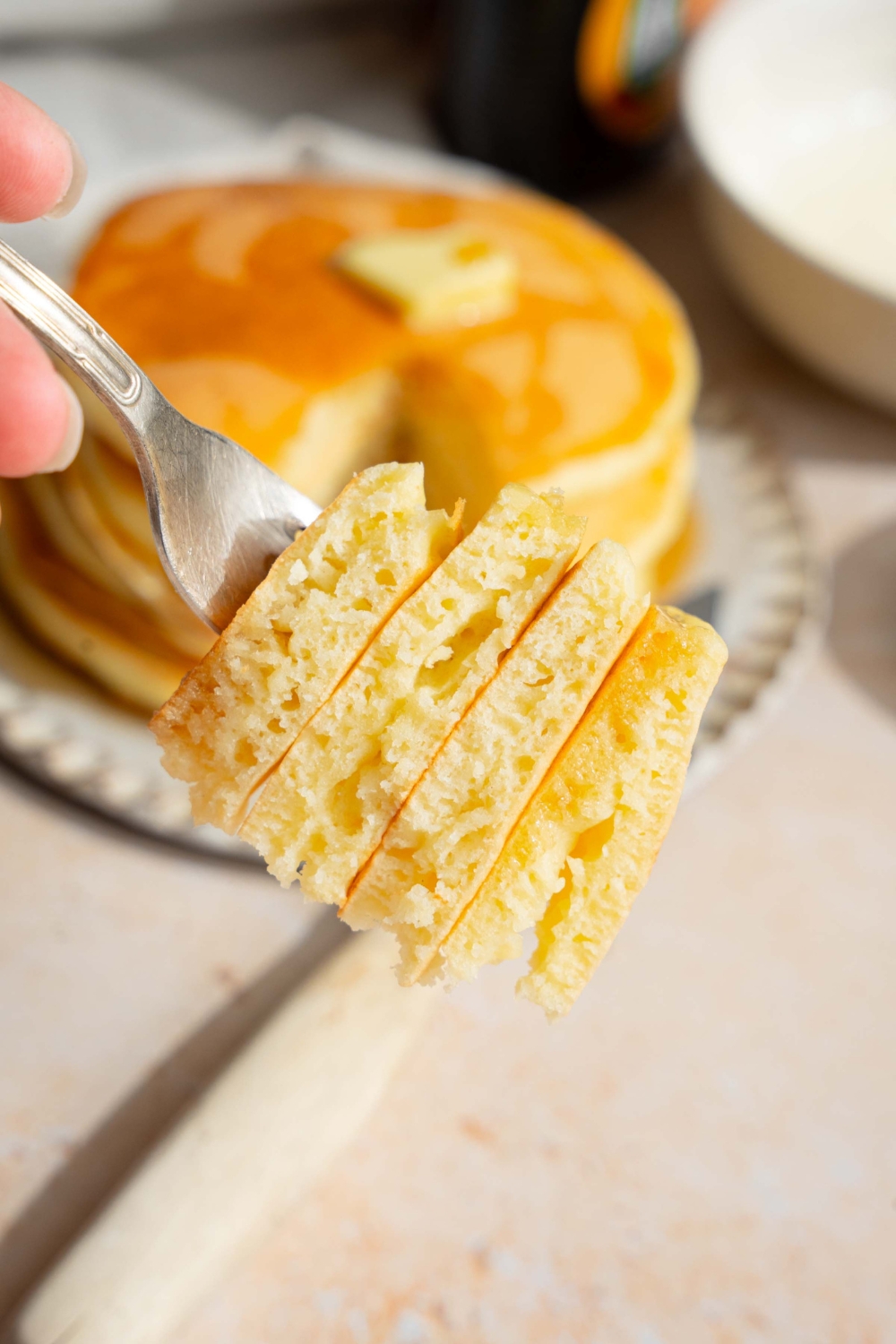 A close up of a fork with a bite of pancakes with self rising flour taken from the stack. There is a plate of pancakes on a tan counter blurred in the background.