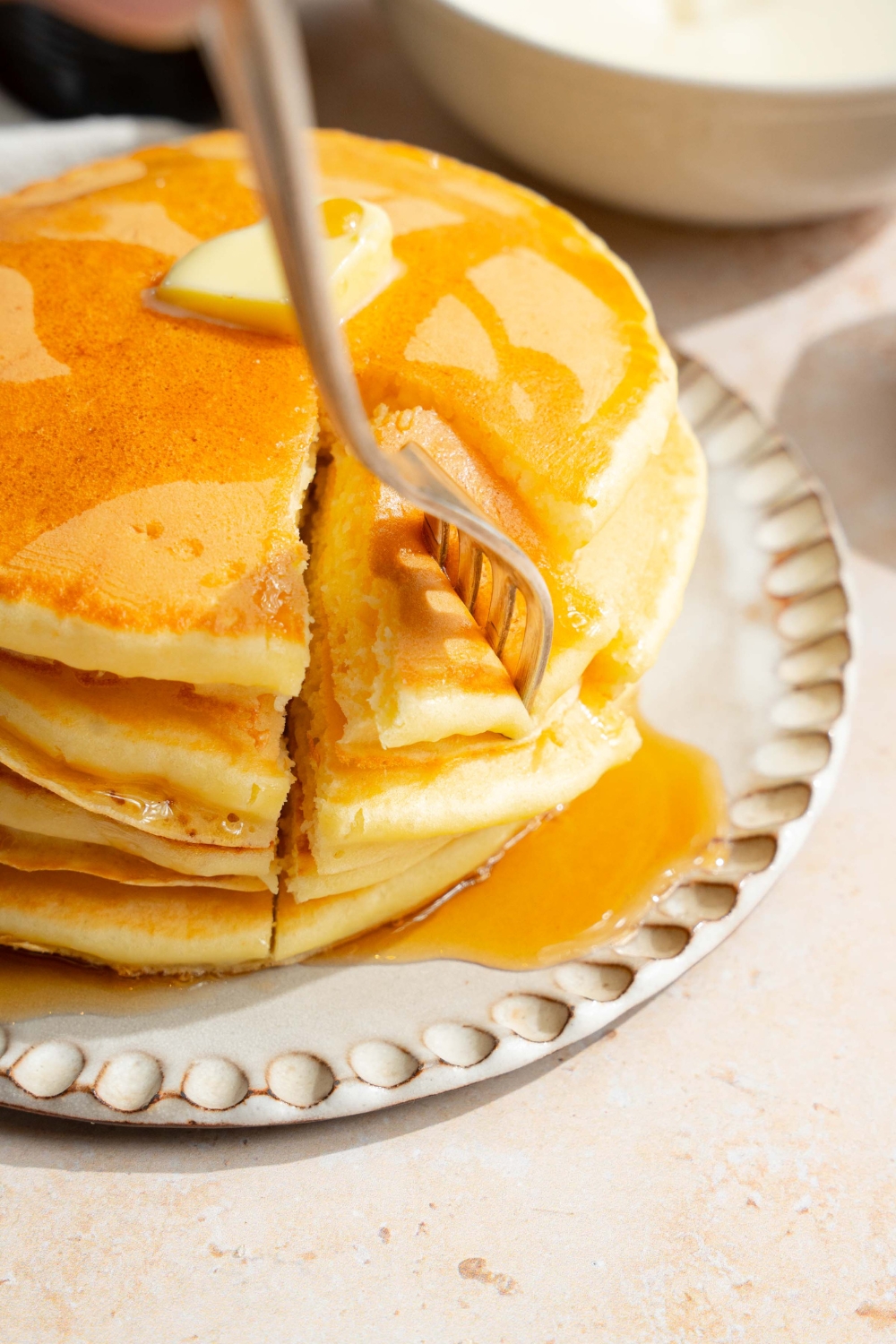A white plate with a stack of pancakes with self rising flour topped with a pat of butter and drizzled with syrup. A fork is taking a bite from the stack. The plate is on a tan counter.