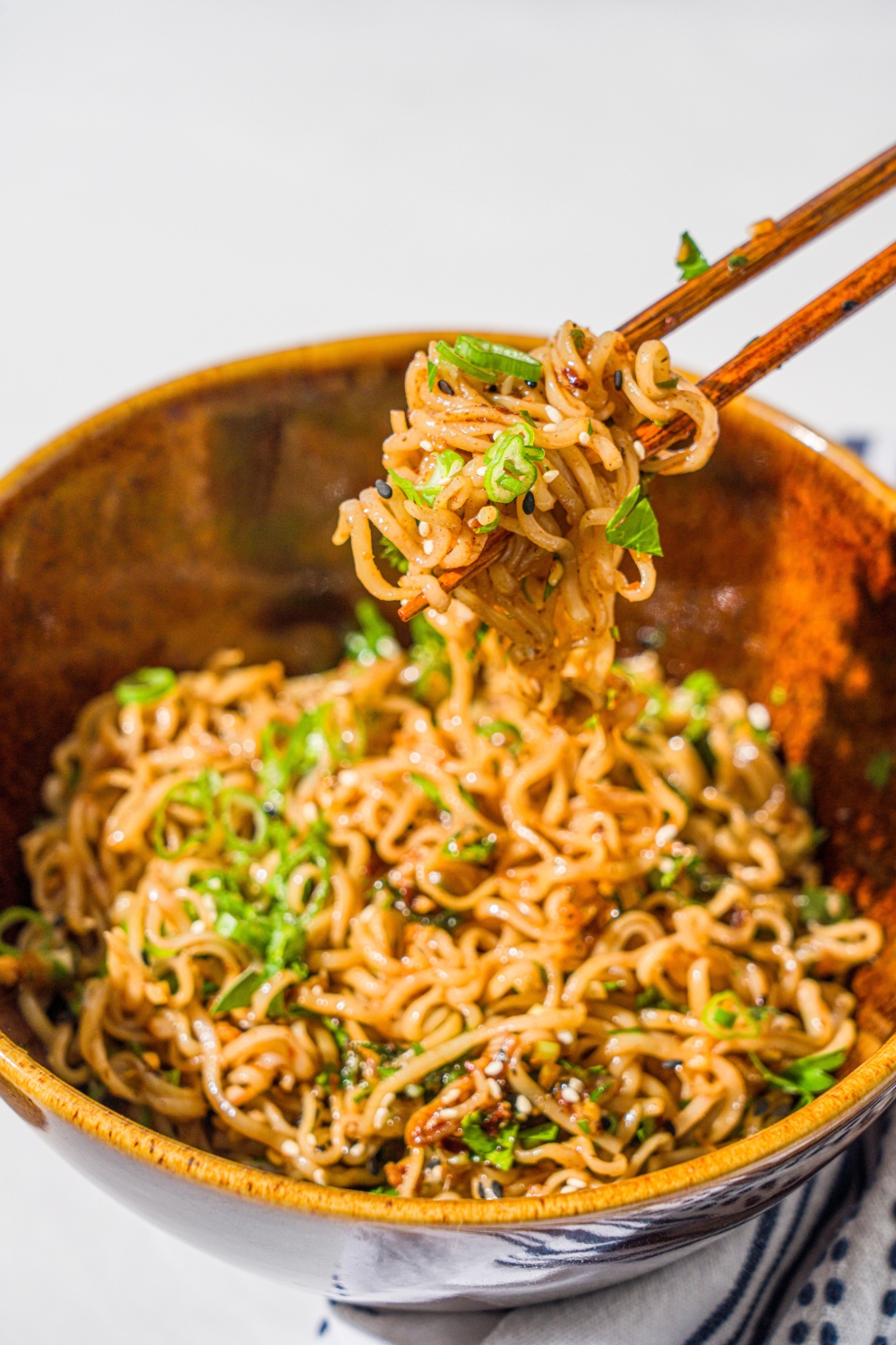 A ceramic bowl with garlic chili oil noodles garnished with sliced green onion and sesame seeds. There is a pair of chopsticks taking a bite of noodles from the bowl. The bowl is on a marble counter with a white cloth napkin.