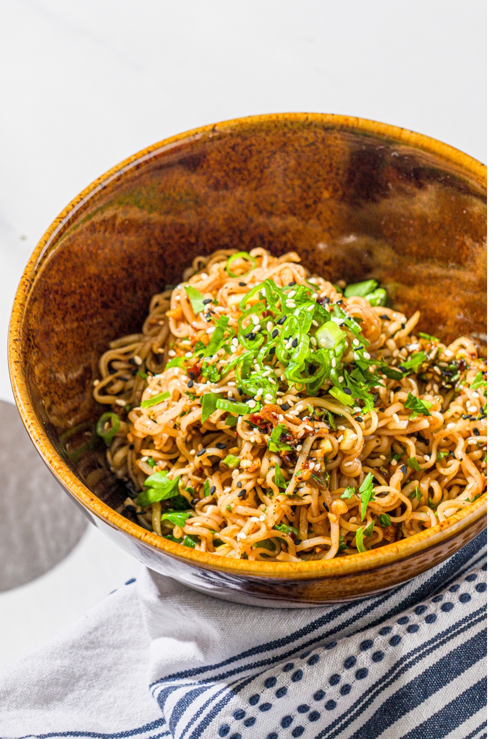 A ceramic bowl with garlic chili oil noodles garnished with sliced green onion and sesame seeds. The bowl is on a marble counter with a white cloth napkin.