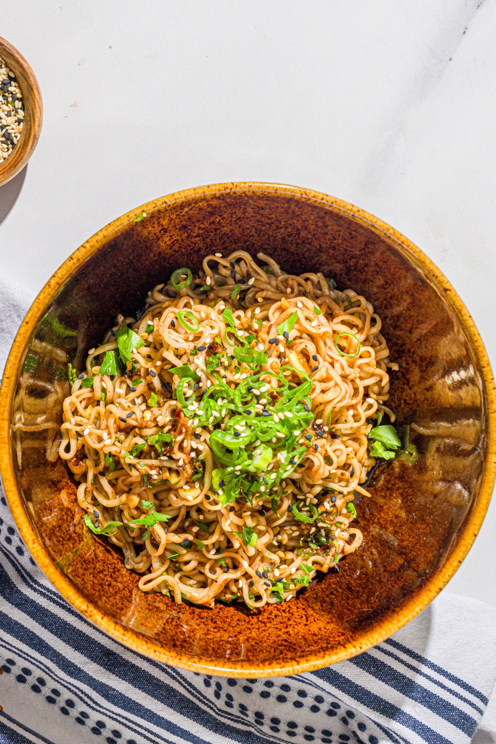 A ceramic bowl with garlic chili oil noodles garnished with sliced green onion and sesame seeds. The bowl is on a marble counter with a white cloth napkin.