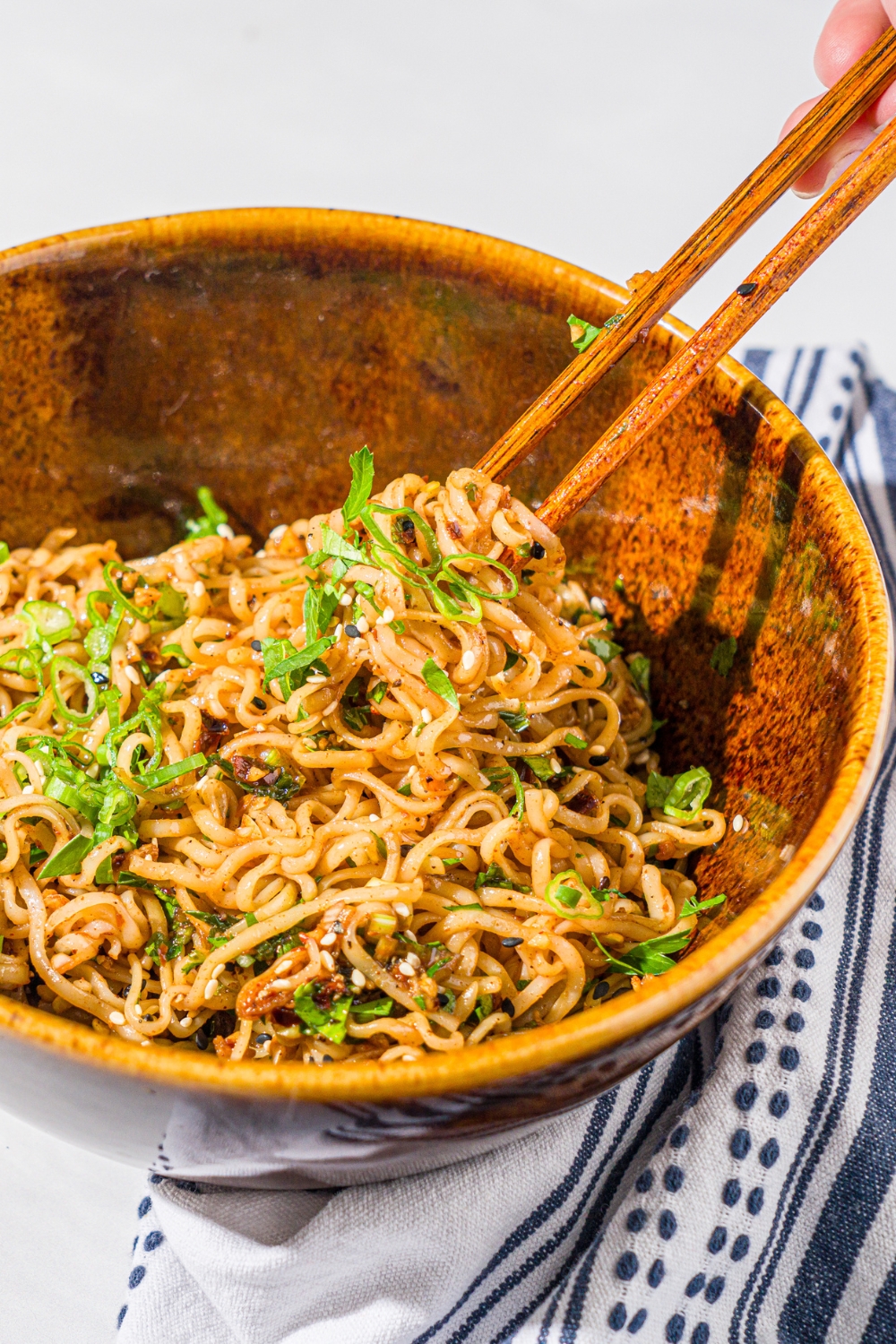 A ceramic bowl with garlic chili oil noodles garnished with sliced green onion and sesame seeds. A pair of chopsticks is taking a bite of noodles from the bowl. The bowl is on a marble counter with a white cloth napkin.