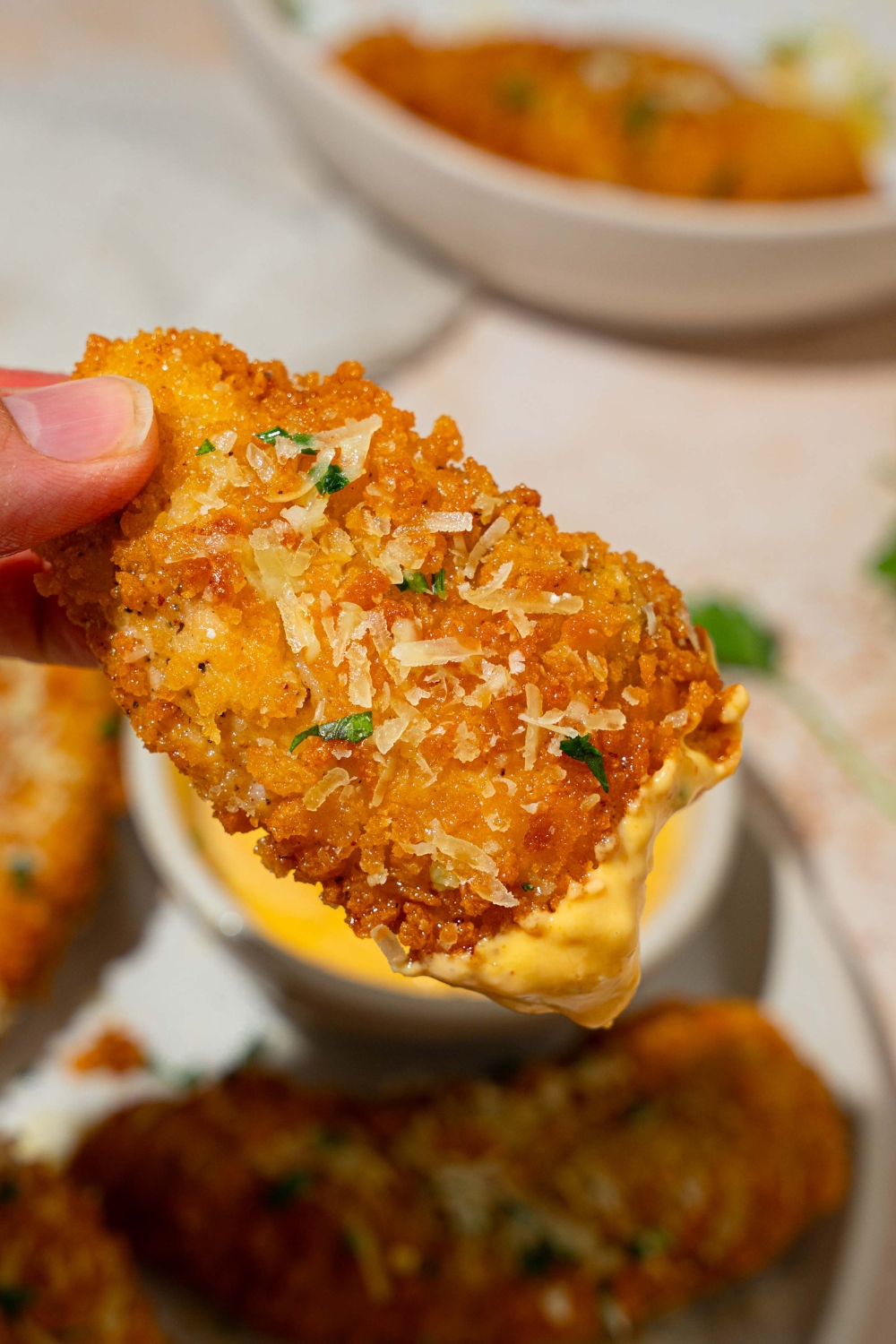 A close up of a garlic parmesan chicken tender garnished with shredded parmesan cheese and fresh parsley dipped in sauce. There is a white plate of chicken tenders on a tan counter blurred in the background.