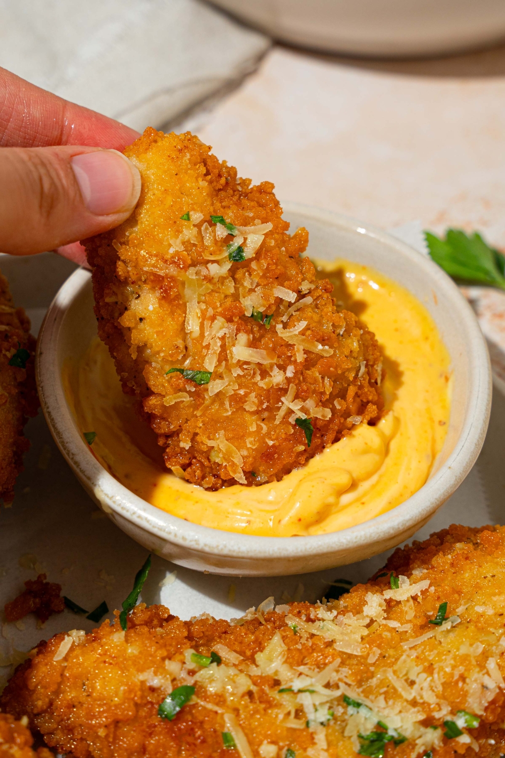 A hand dipping a garlic parmesan chicken tender into a bowl of dip. The bowl is on a white plate with chicken tenders garnished with shredded parmesan cheese and fresh parsley. The plate is on a tan counter with a white cloth napkin.
