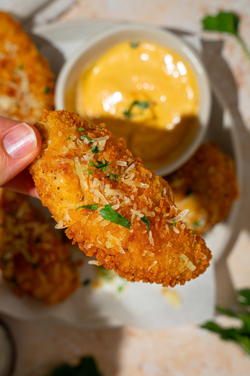 A close up of a garlic parmesan chicken tender garnished with shredded parmesan cheese and fresh parsley. There is a white plate of chicken tenders on a tan counter blurred in the background.