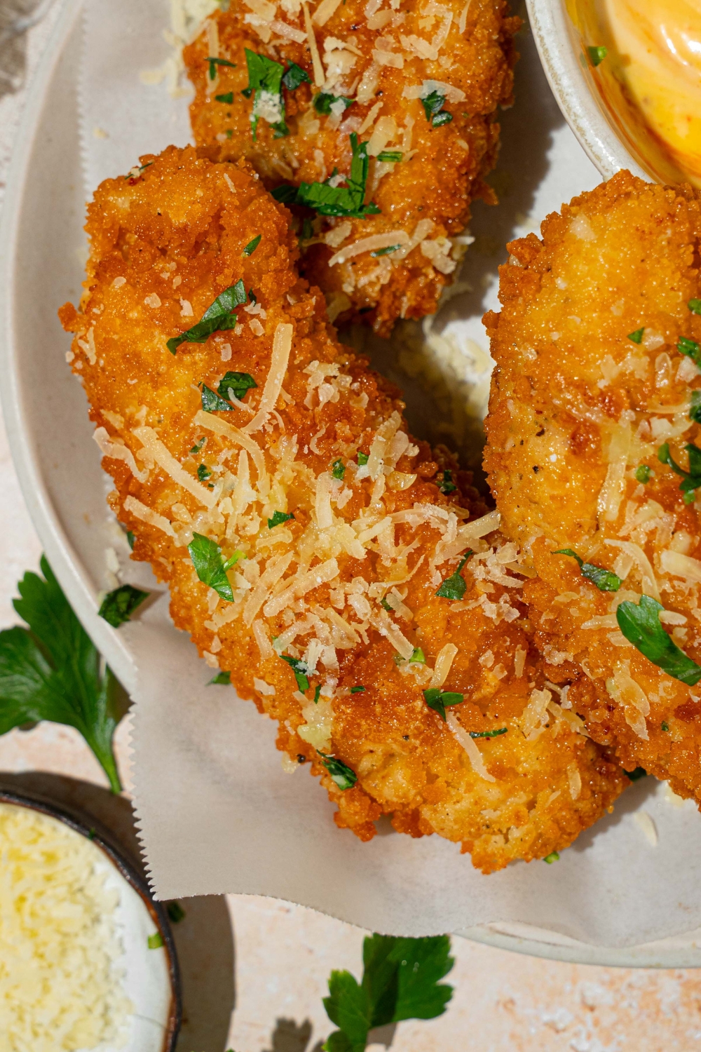 A white plate lined with parchment paper with garlic parmesan chicken tenders garnished with parmesan cheese and fresh parsley. There is a bowl of dip on the plate. The plate is on a tan counter.