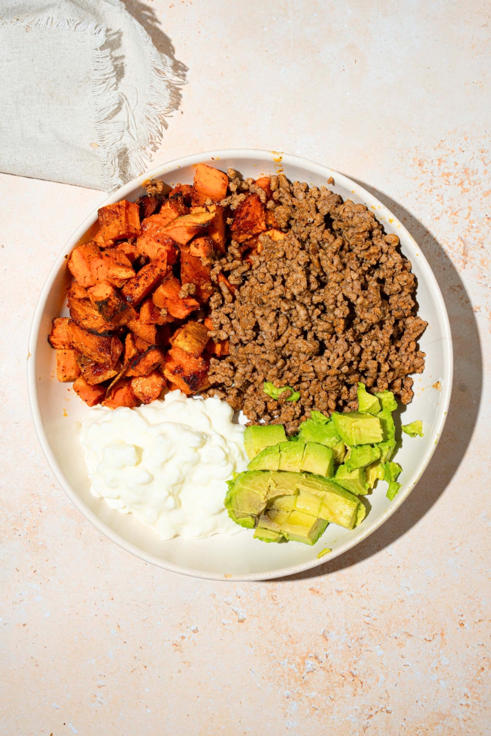 Diced roasted sweet potatoes, ground beef, sliced avocado, and cottage cheese on a white plate on a white counter.