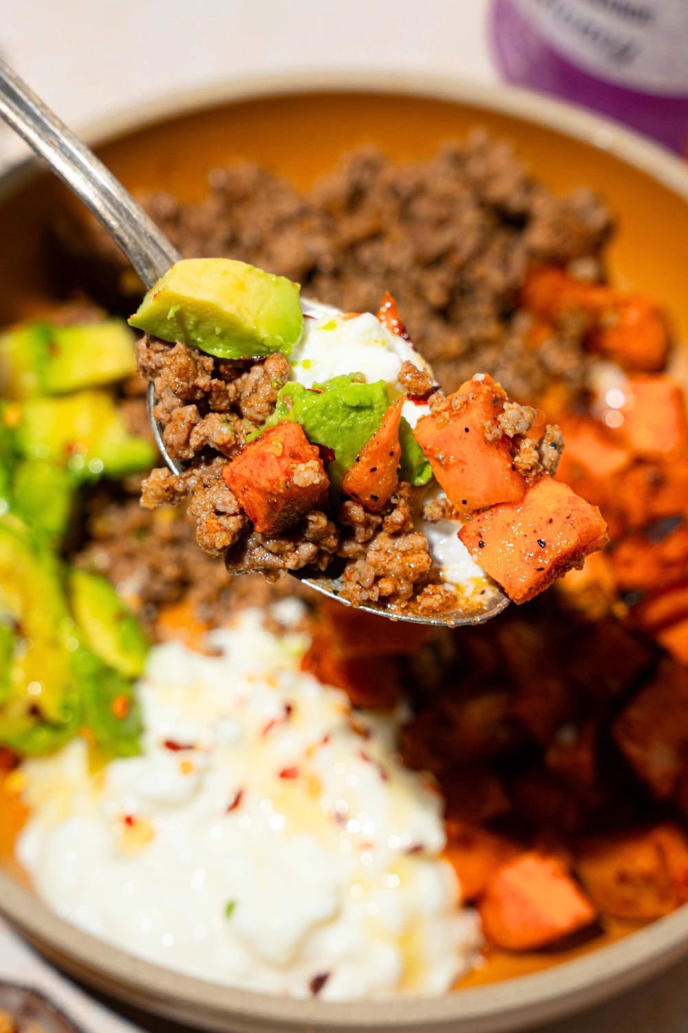 A spoon with some avocado, ground beef, diced, roasted sweet potatoes, and cottage cheese being held above a ground beef, sweet potato cottage cheese bowl.