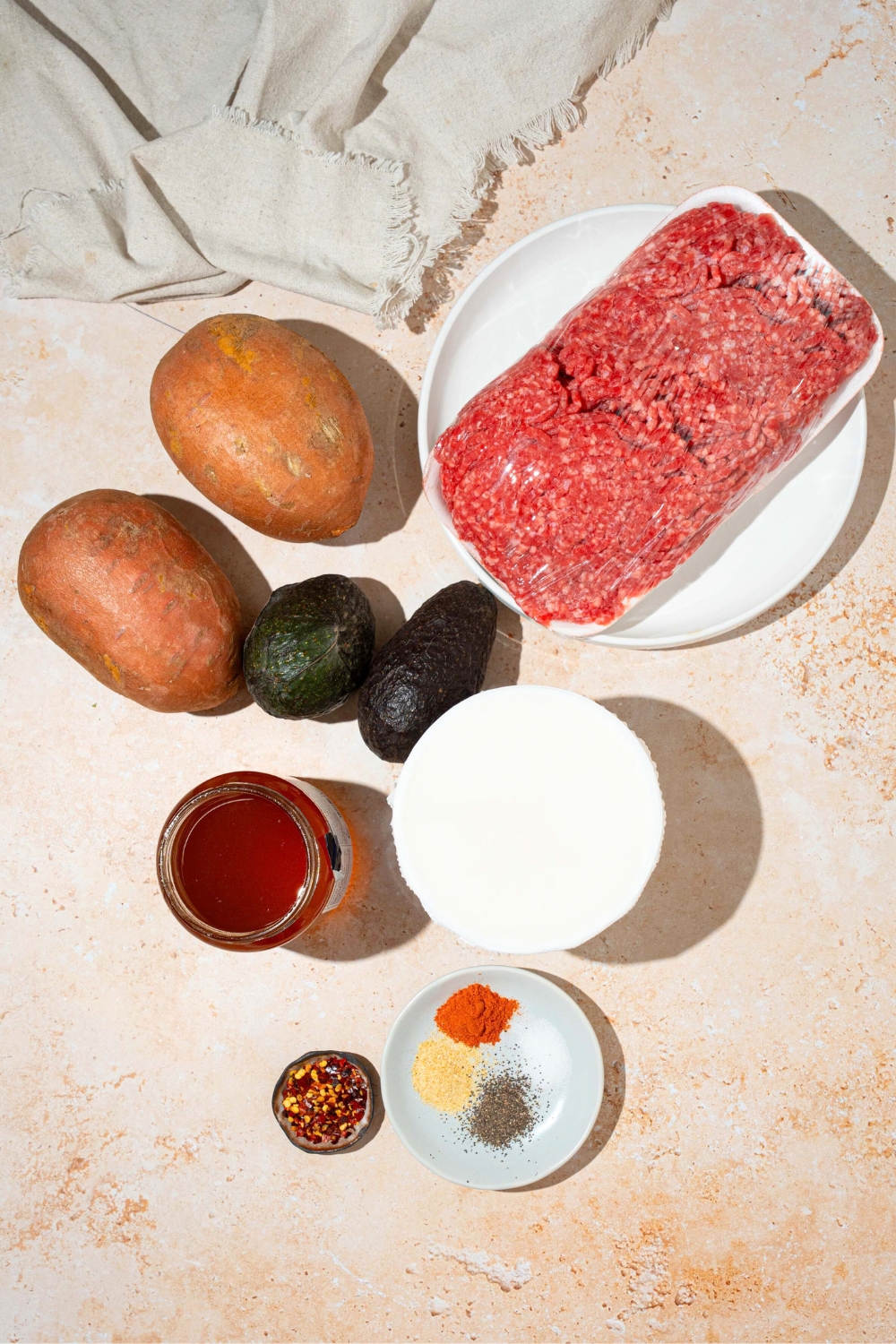 A pack of ground beef, two sweet potatoes, two avocados, a bowl of hot honey, a bowl of cottage cheese, a small plate with seasoning on it, and a small dish of red pepper flakes all on a white counter.