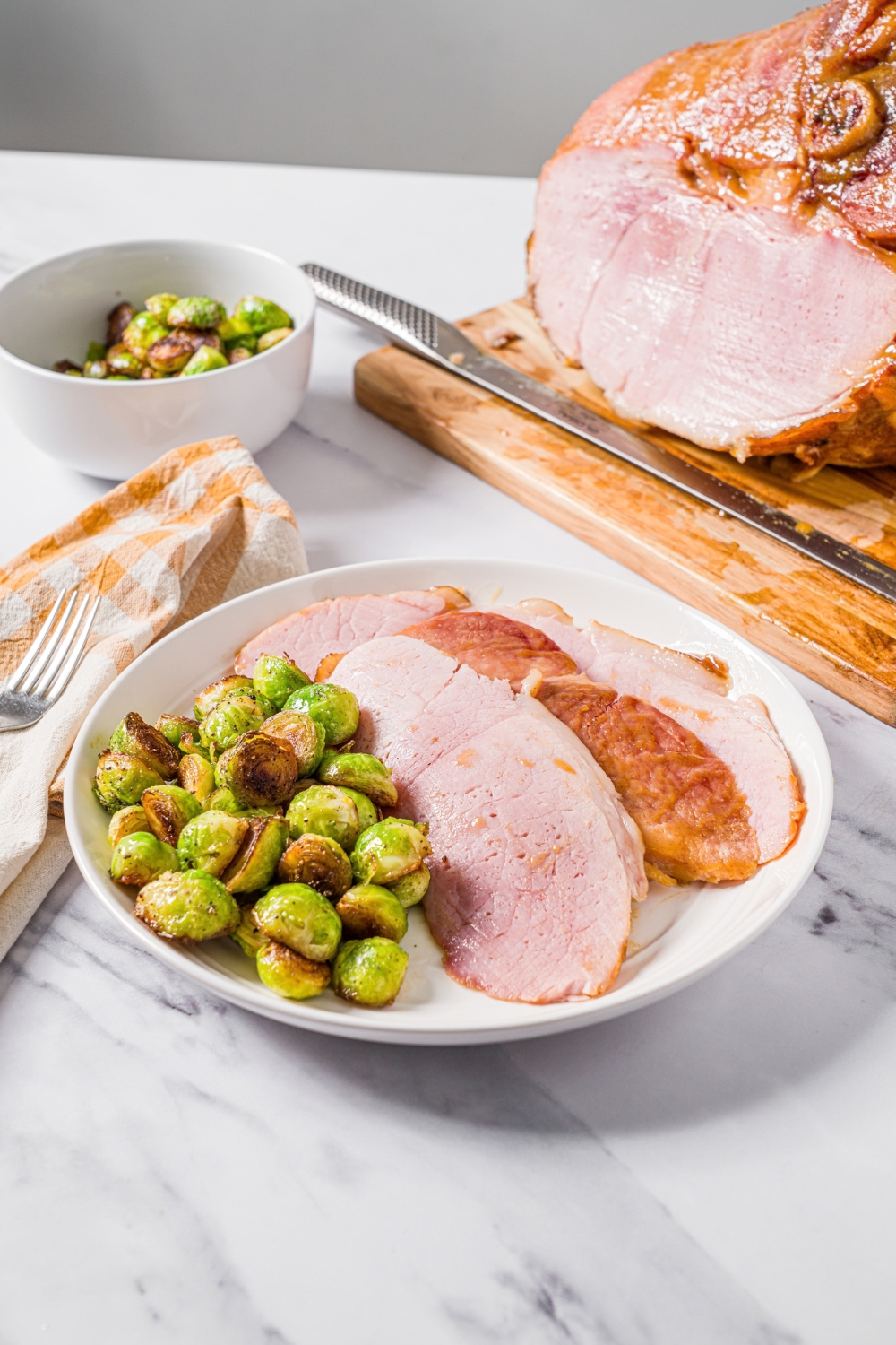 Ham shank slices on a white plate with some brussels sprouts. Behind that is a whole ham shank on a wooden cutting board and a white bowl filled with brussels sprouts.