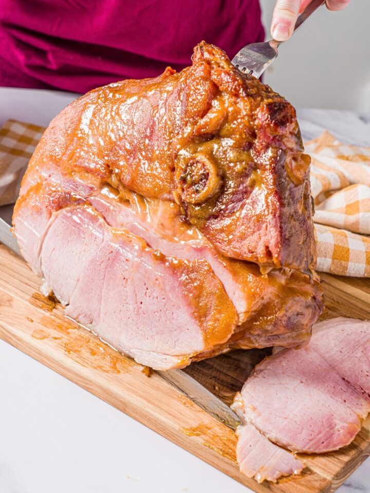 Two hands slicing a glazed ham shank that is on a wooden cutting board.