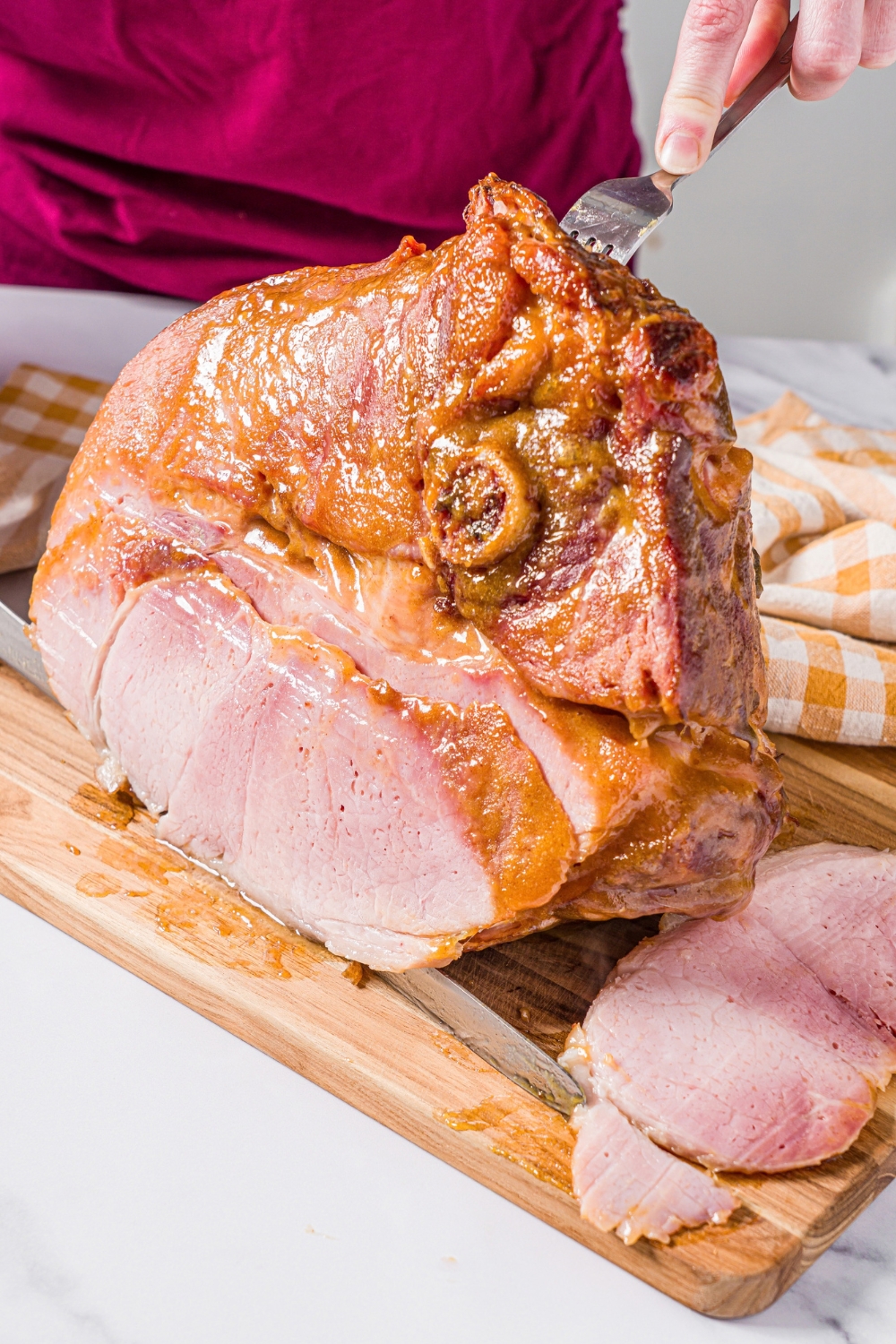 Two hands slicing a glazed ham shank that is on a wooden cutting board.