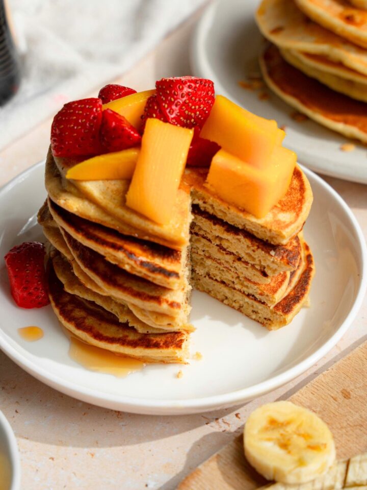A stack of banana oat pancakes that have a triangle slice out of the front of the stack. There are sliced strawberries and mangoes on top of the stack of pancakes.