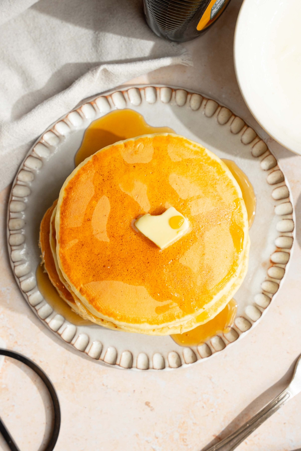 A white plate with a stack of pancakes with self rising flour topped with a pat of butter and drizzled with syrup. The plate is on a tan counter with a white cloth napkin.
