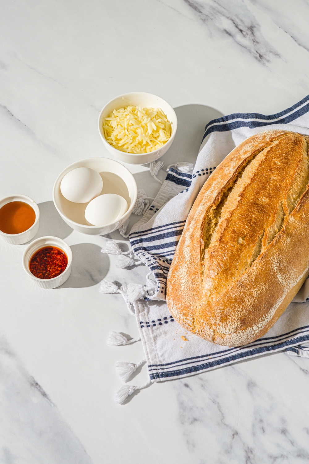 A loaf of sourdough, a bowl of two eggs, a bowl of honey, a bowl of chili crisps, and a bowl of monterey jack cheese all on a white counter. The sourdough loaf is on a blue and white towel on the counter.