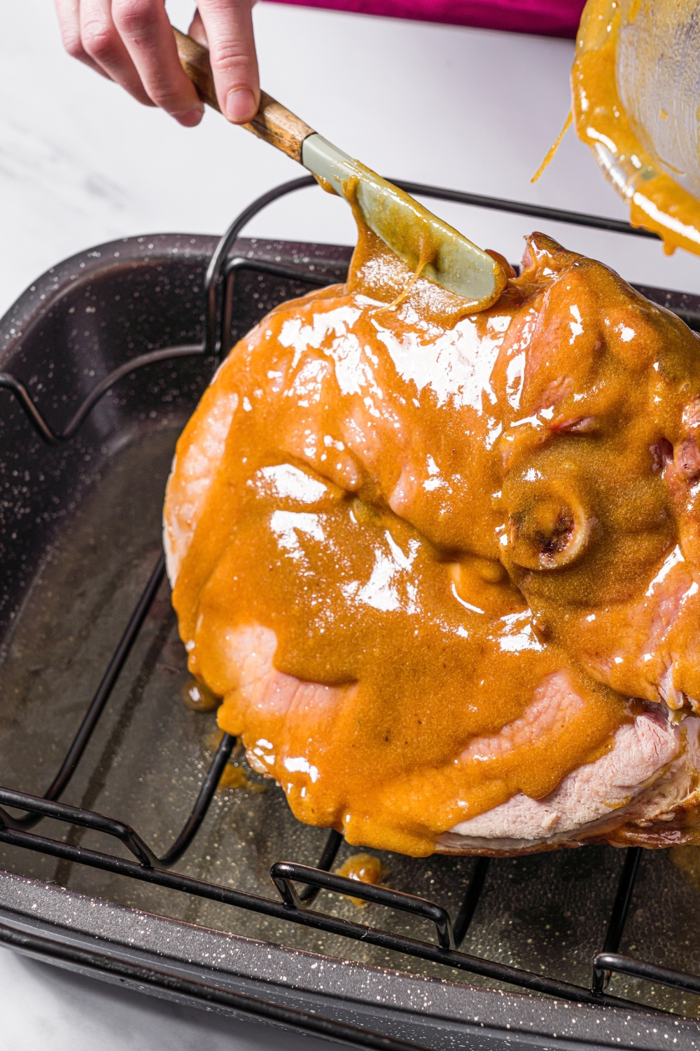 A hand brushing a brown sugar mustard glaze onto a ham shank in a roasting pan.