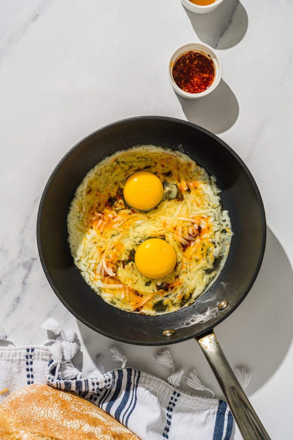 Two eggs on top of shredded cheese and chili crisps cooking in a skillet to make hot honey eggs.