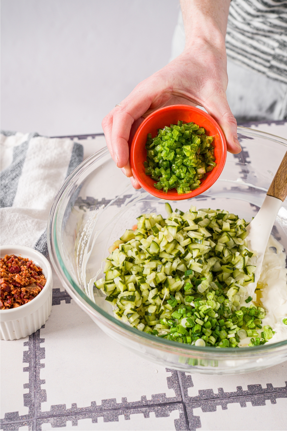 A hand pouring diced jalapeños into a glass bowl that has chopped pickles and a creamy mixture in it.