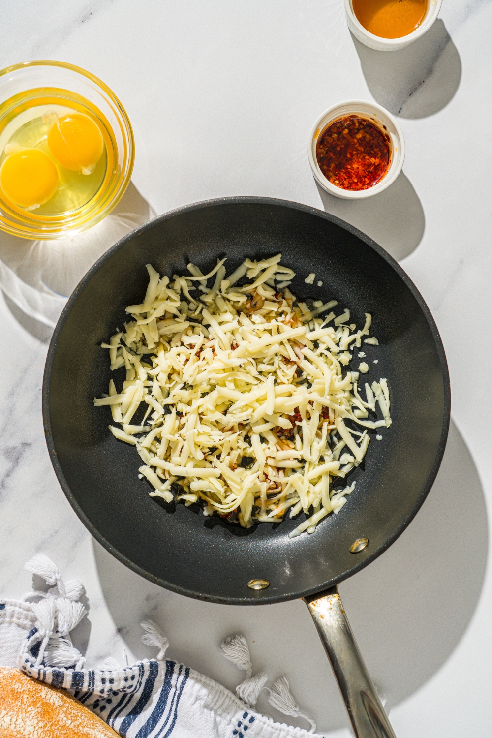 Shredded monterey jack cheese with honey drizzled on top of it in a skillet. There is small bowl of two eggs and a small bowl of chili crisp in front of it to make hot honey eggs.