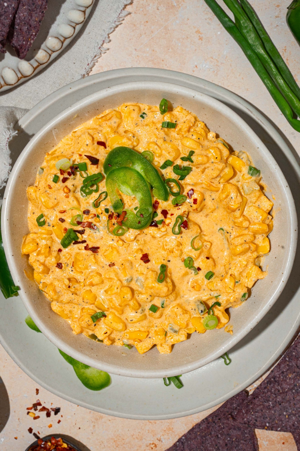A bowl of jalapeño corn dip garnished with sliced green onion and sliced jalapeños. The bowl is on served on a white plate with sliced jalapeños. The plate is on a tan counter with blue corn chips.