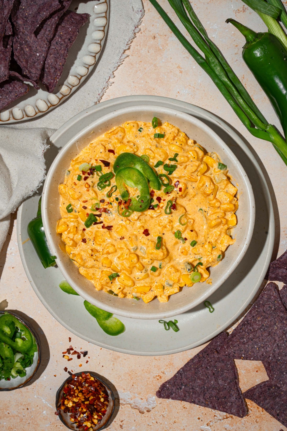 A bowl of jalapeño corn dip garnished with sliced green onion and sliced jalapeños. The bowl is on served on a white plate with sliced jalapeños. The plate is on a tan counter with blue corn chips and small bowls of garnishes.