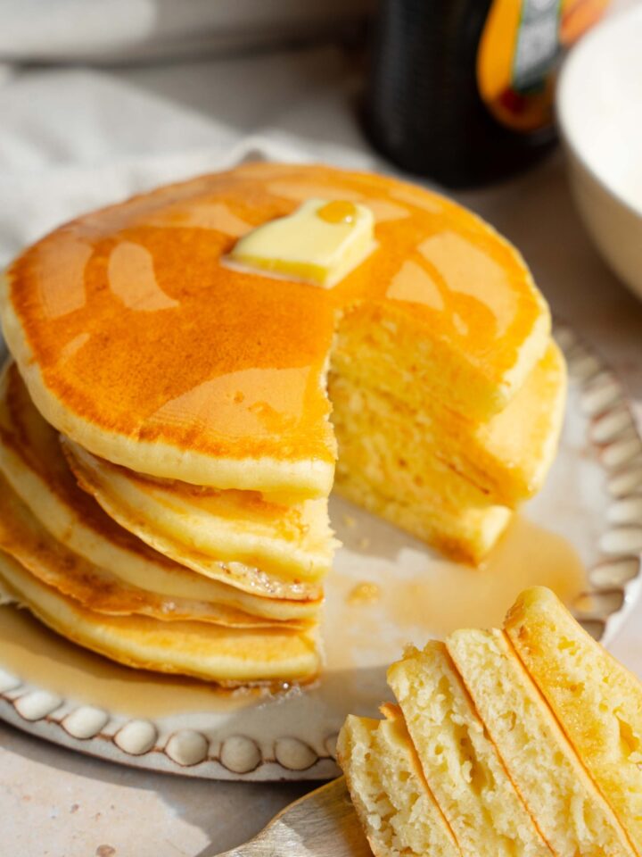 A white plate with a stack of pancakes with self rising flour topped with a pat of butter and drizzled with syrup. A fork is taking a bite from the stack. The plate is on a tan counter with a jar of syrup.