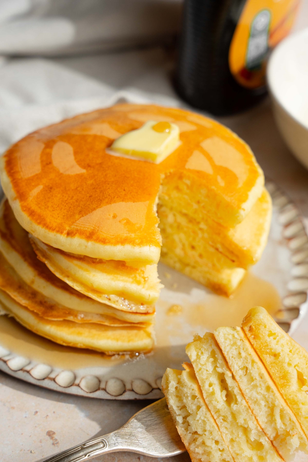 A white plate with a stack of pancakes with self rising flour topped with a pat of butter and drizzled with syrup. A fork is taking a bite from the stack. The plate is on a tan counter with a jar of syrup.