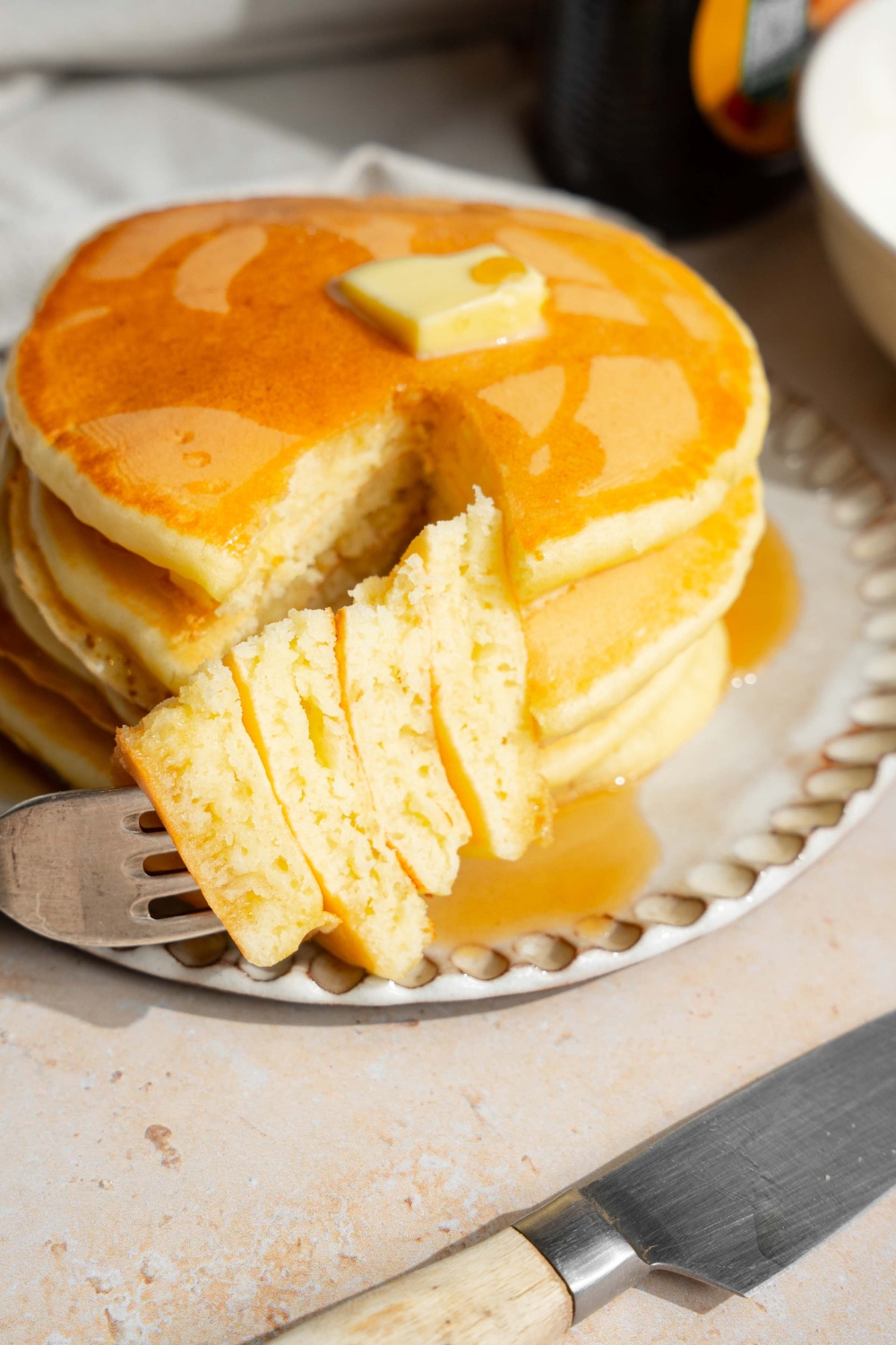 A white plate with a stack of pancakes with self rising flour topped with a pat of butter and drizzled with syrup. A fork is taking a bite from the stack. The plate is on a tan counter with a jar of syrup and a knife.
