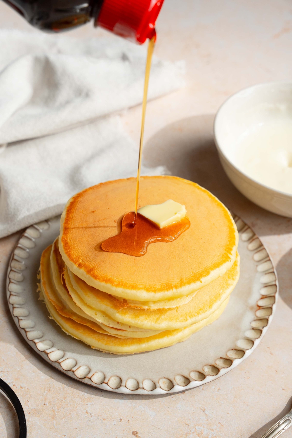 A white plate with a stack of pancakes with self rising flour topped with a pat of butter. Syrup is being poured over the stack. The plate is on a tan counter with a white cloth napkin.