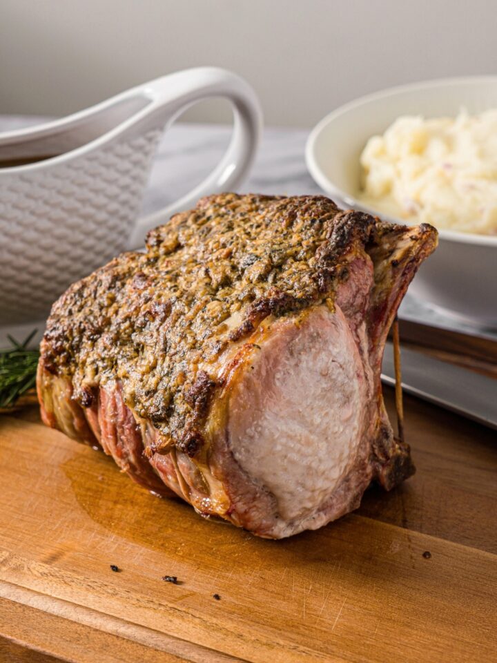 A pork rib roast on a wooden cutting board with part of a bowl of mashed potatoes and part of a gravy boat behind it.