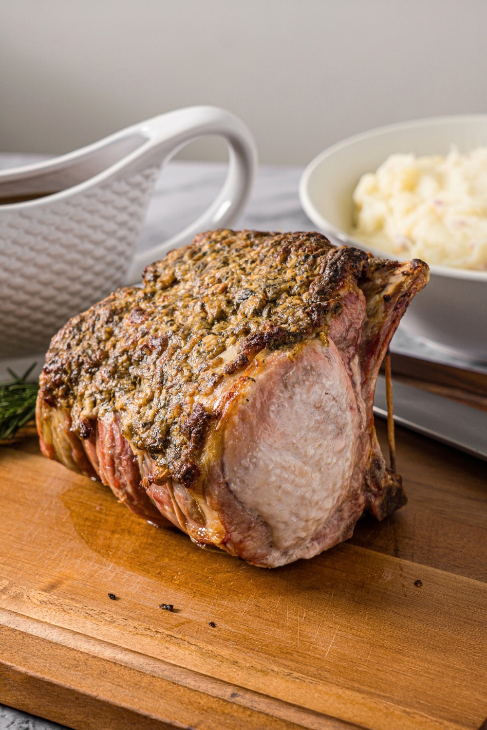 A pork rib roast on a wooden cutting board with part of a bowl of mashed potatoes and part of a gravy boat behind it.