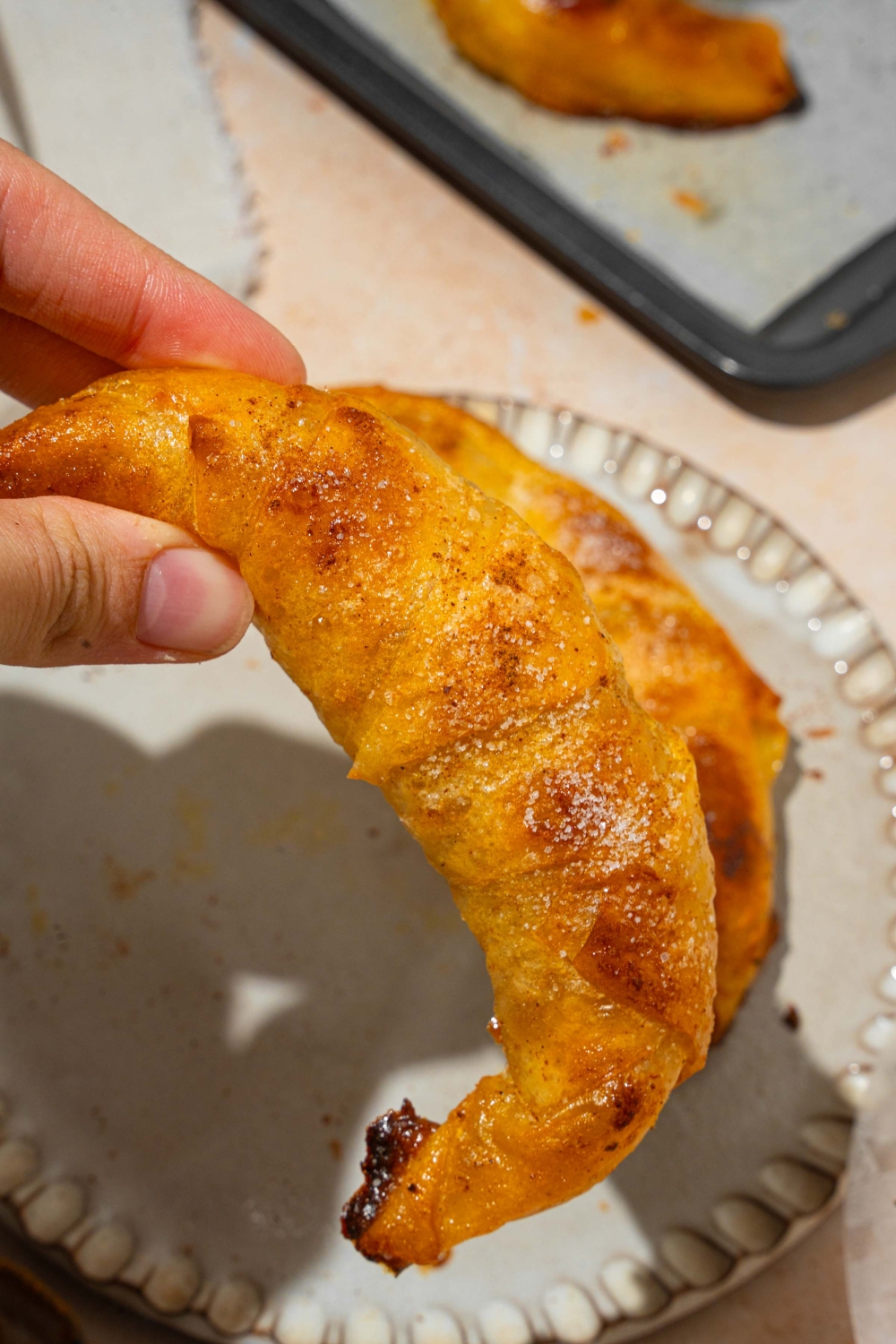 A close up of a hand holding a rice paper croissant. There is a plate with a croissant on a tan counter with a baking sheet of croissants blurred in the background.
