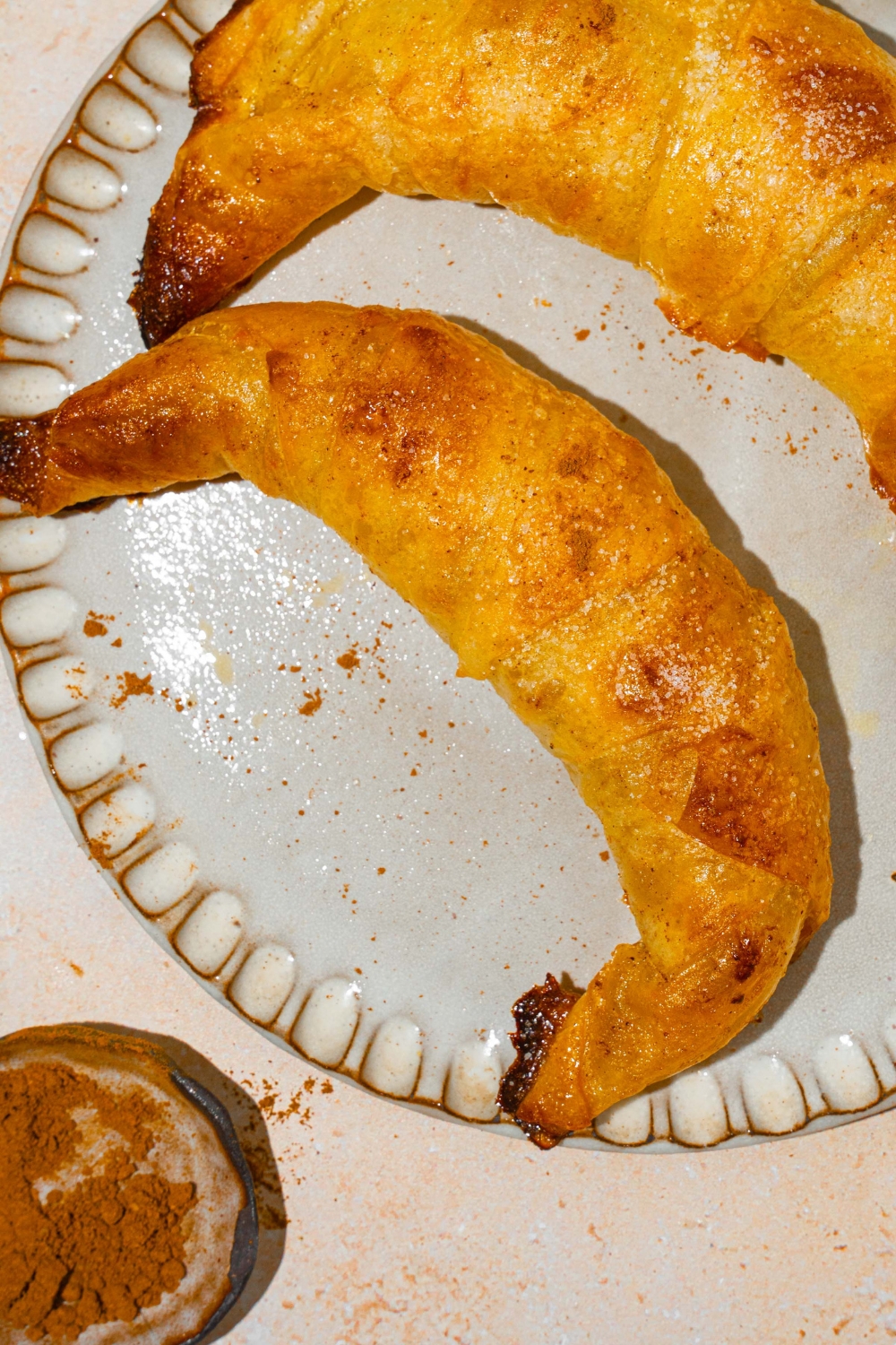 A plate with two baked rice paper croissants. The plate is on a tan counter with a small bowl of cinnamon.