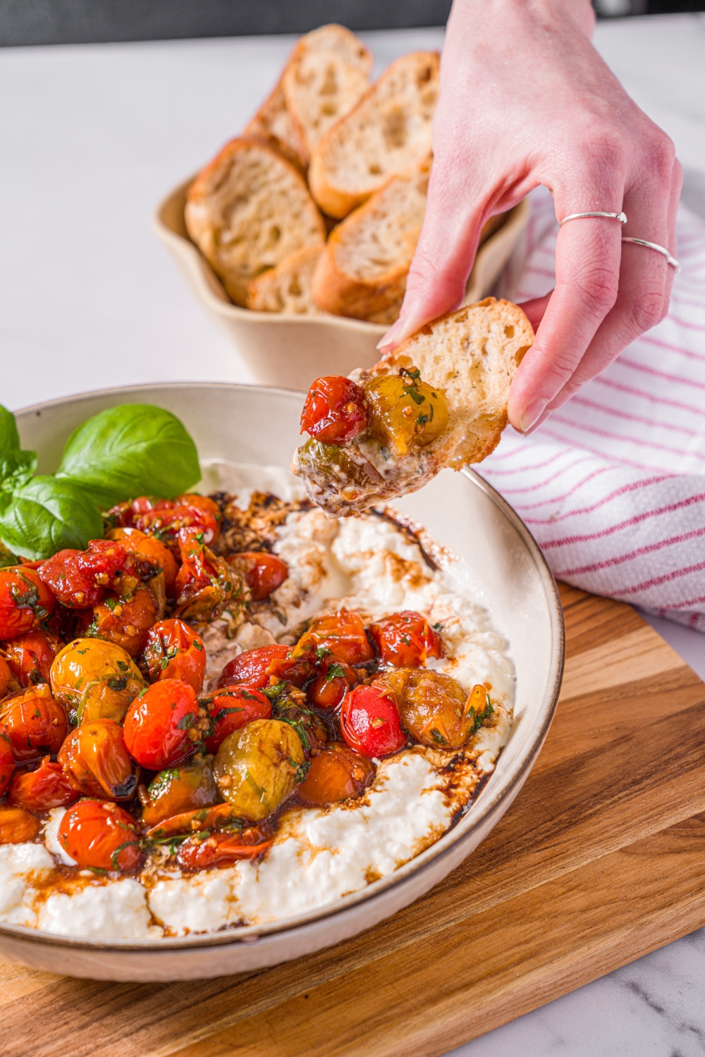 A white bowl with roasted tomato burrata dip garnished with fresh basil. A hand is dipping a toasted crostini in the dip. The bowl is on a wooden board on a marble counter with a small bowl of toasted crostini.