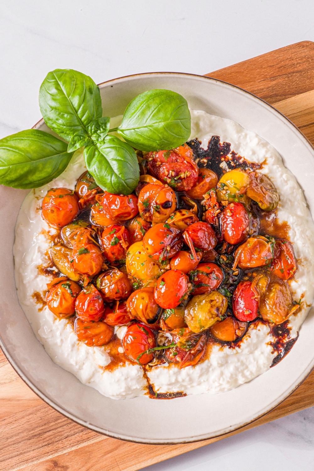 A white bowl with roasted tomato burrata dip garnished with fresh basil. The bowl is on a wooden board on a marble counter.